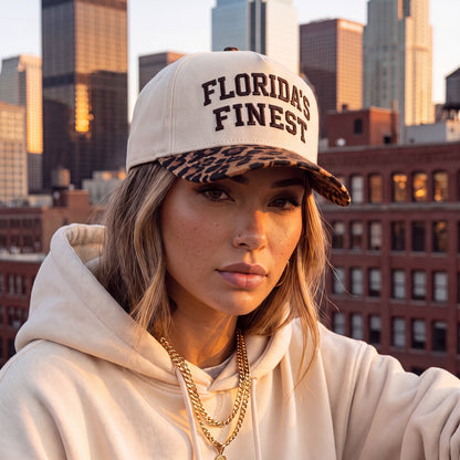 Woman wearing a cream Florida’s Finest hat with a leopard-print brim and matching cream hoodie, posed against a city skyline at sunset.