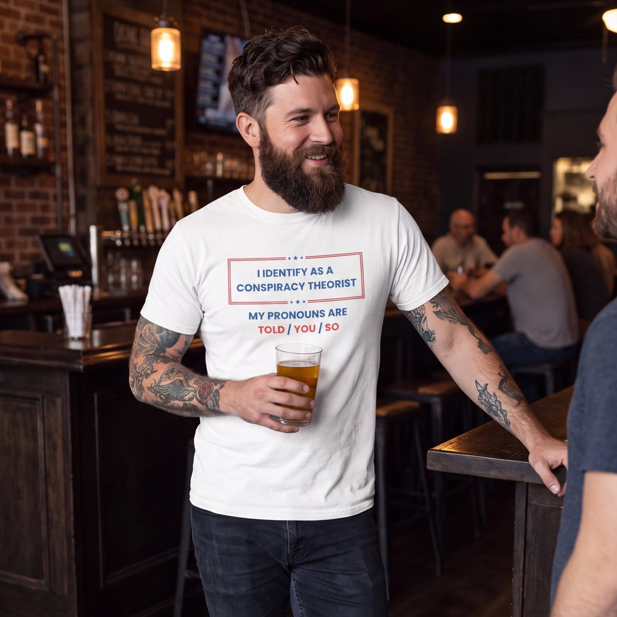 Bearded man wearing a white graphic t-shirt reading “I Identify as a Conspiracy Theorist, My Pronouns Are Told/You/So,” holding a drink while talking with a friend at a bar.