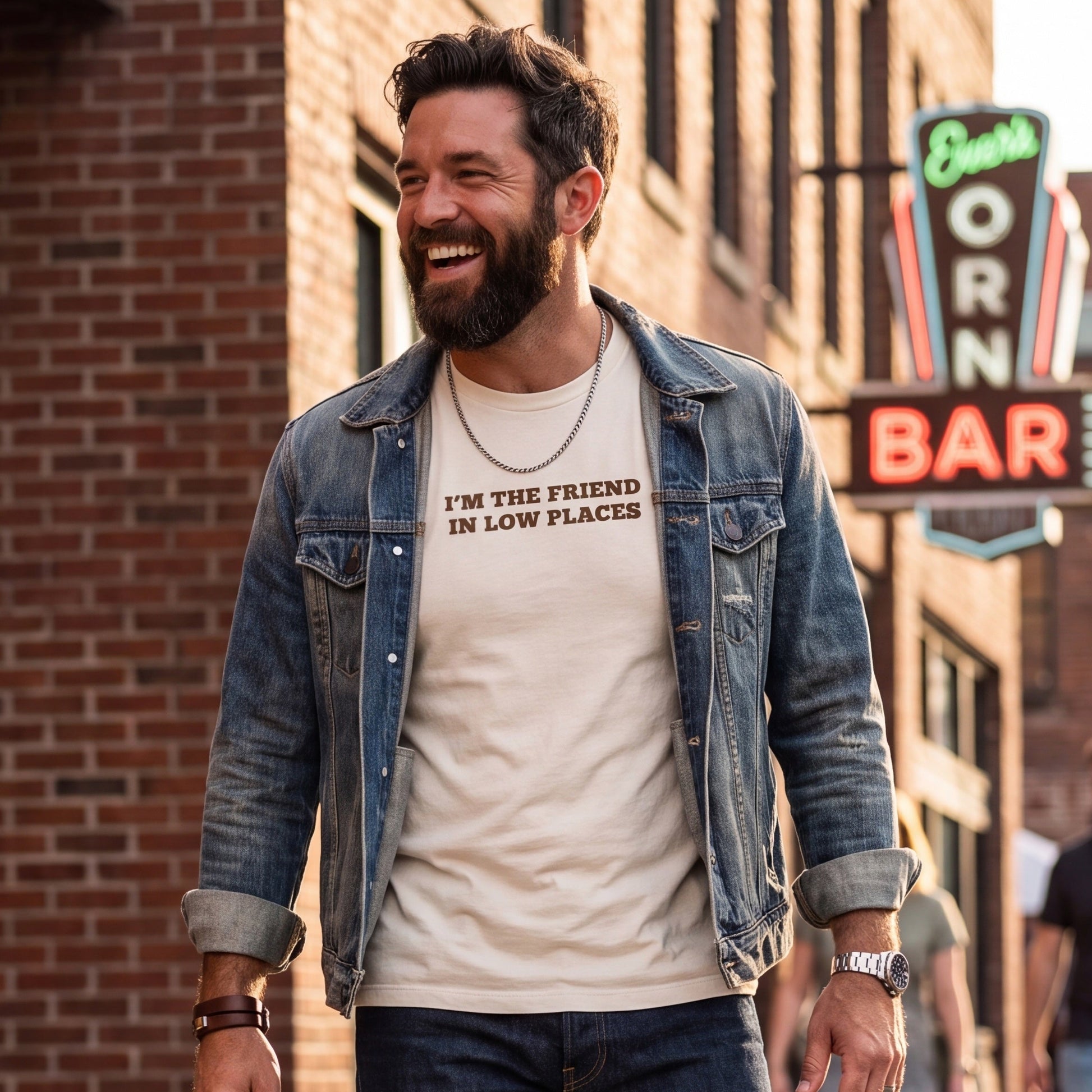 Smiling bearded man wearing a cream graphic t-shirt with brown text reading “I’M THE FRIEND IN LOW PLACES,” layered under a denim jacket. He walks through a brick-lined entertainment district near a glowing bar sign, creating a casual lifestyle setting for the humorous statement tee.