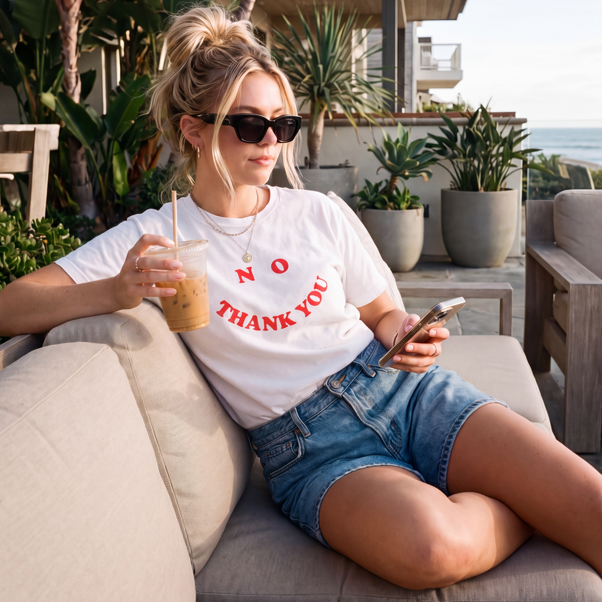 A woman sits on a beige outdoor sofa wearing a white graphic T-shirt that reads 'NO THANK YOU' in red letters, blue denim shorts, and black sunglasses while holding an iced coffee and phone.