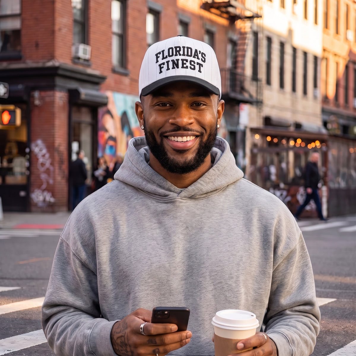 Smiling man wearing a white pinstripe Florida’s Finest hat with a black brim and gray hoodie, holding a coffee and phone on a city street.