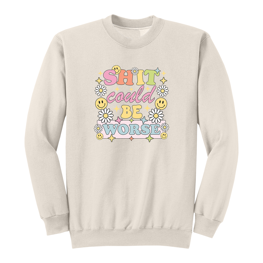 Front view of a natural long sleeve graphic shirt featuring a colorful retro-inspired design with the phrase "SHIT could BE WORSE" in playful pastel typography. The graphic is surrounded by white daisies, yellow smiley faces, and sparkling star accents, creating a cheerful vintage aesthetic that humorously contrasts with the sarcastic message. A funny adult long sleeve shirt with retro floral graphics, bold humor, and casual streetwear style.