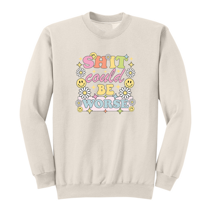 Front view of a natural long sleeve graphic shirt featuring a colorful retro-inspired design with the phrase "SHIT could BE WORSE" in playful pastel typography. The graphic is surrounded by white daisies, yellow smiley faces, and sparkling star accents, creating a cheerful vintage aesthetic that humorously contrasts with the sarcastic message. A funny adult long sleeve shirt with retro floral graphics, bold humor, and casual streetwear style.