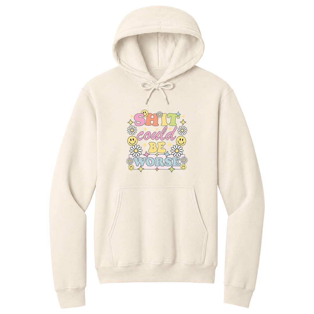 Front view of a natural pullover hoodie featuring a colorful retro-inspired graphic with the phrase "SHIT could BE WORSE" in playful pastel typography. The design is surrounded by white daisies, yellow smiley faces, and sparkling star accents, creating a cheerful vintage aesthetic that humorously contrasts with the sarcastic message. The drawstrings are tied into a short bow above the design for a realistic product mockup. A funny adult hoodie with bold humor, retro floral graphics, and streetwear style