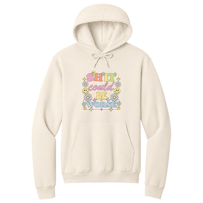 Front view of a natural pullover hoodie featuring a colorful retro-inspired graphic with the phrase "SHIT could BE WORSE" in playful pastel typography. The design is surrounded by white daisies, yellow smiley faces, and sparkling star accents, creating a cheerful vintage aesthetic that humorously contrasts with the sarcastic message. The drawstrings are tied into a short bow above the design for a realistic product mockup. A funny adult hoodie with bold humor, retro floral graphics, and streetwear style