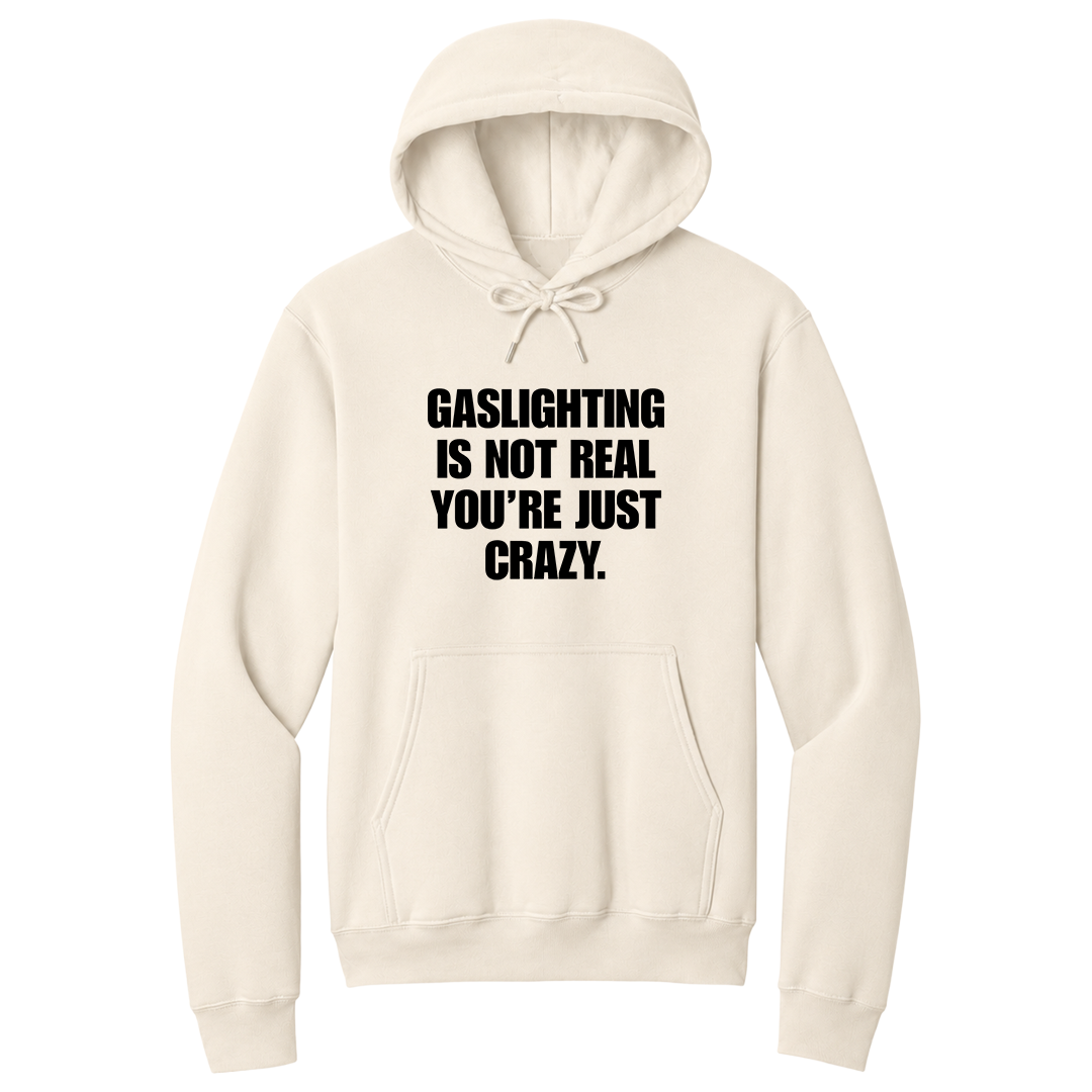 Front view of a natural pullover hoodie featuring the phrase "GASLIGHTING IS NOT REAL YOU’RE JUST CRAZY." printed in bold black uppercase lettering centered on the chest. The hoodie includes a front kangaroo pocket and matching black drawstrings tied in a bow, with the minimalist black-and-white design creating a bold, sarcastic statement look.