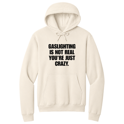 Front view of a natural pullover hoodie featuring the phrase "GASLIGHTING IS NOT REAL YOU’RE JUST CRAZY." printed in bold black uppercase lettering centered on the chest. The hoodie includes a front kangaroo pocket and matching black drawstrings tied in a bow, with the minimalist black-and-white design creating a bold, sarcastic statement look.