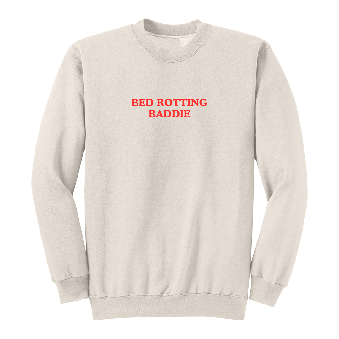 Front view of a natural crewneck sweatshirt featuring the phrase "BED ROTTING BADDIE" in small vintage-style red lettering centered on the chest. This minimalist graphic sweatshirt combines cozy comfort with trendy Gen Z-inspired humor, making it perfect for lounging, casual outings, and everyday wear.