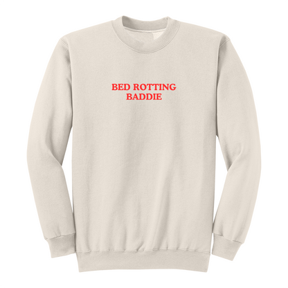 Front view of a natural crewneck sweatshirt featuring the phrase "BED ROTTING BADDIE" in small vintage-style red lettering centered on the chest. This minimalist graphic sweatshirt combines cozy comfort with trendy Gen Z-inspired humor, making it perfect for lounging, casual outings, and everyday wear.