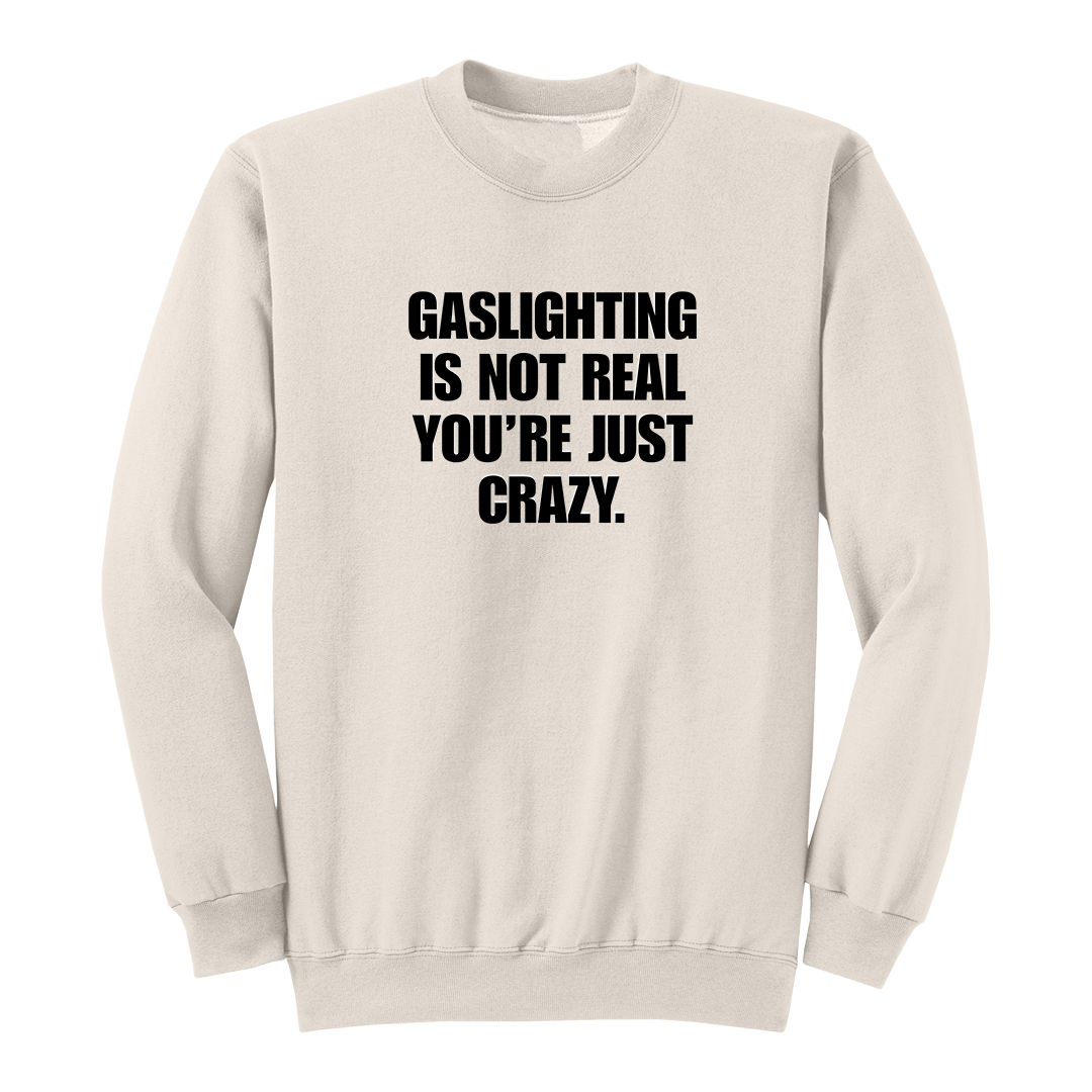 Front view of a natural crewneck sweatshirt featuring the phrase "GASLIGHTING IS NOT REAL YOU’RE JUST CRAZY." printed in bold black uppercase lettering centered on the chest. The clean black-and-white design creates a minimalist statement sweatshirt with a bold, sarcastic aesthetic.