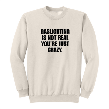 Front view of a natural crewneck sweatshirt featuring the phrase "GASLIGHTING IS NOT REAL YOU’RE JUST CRAZY." printed in bold black uppercase lettering centered on the chest. The clean black-and-white design creates a minimalist statement sweatshirt with a bold, sarcastic aesthetic.