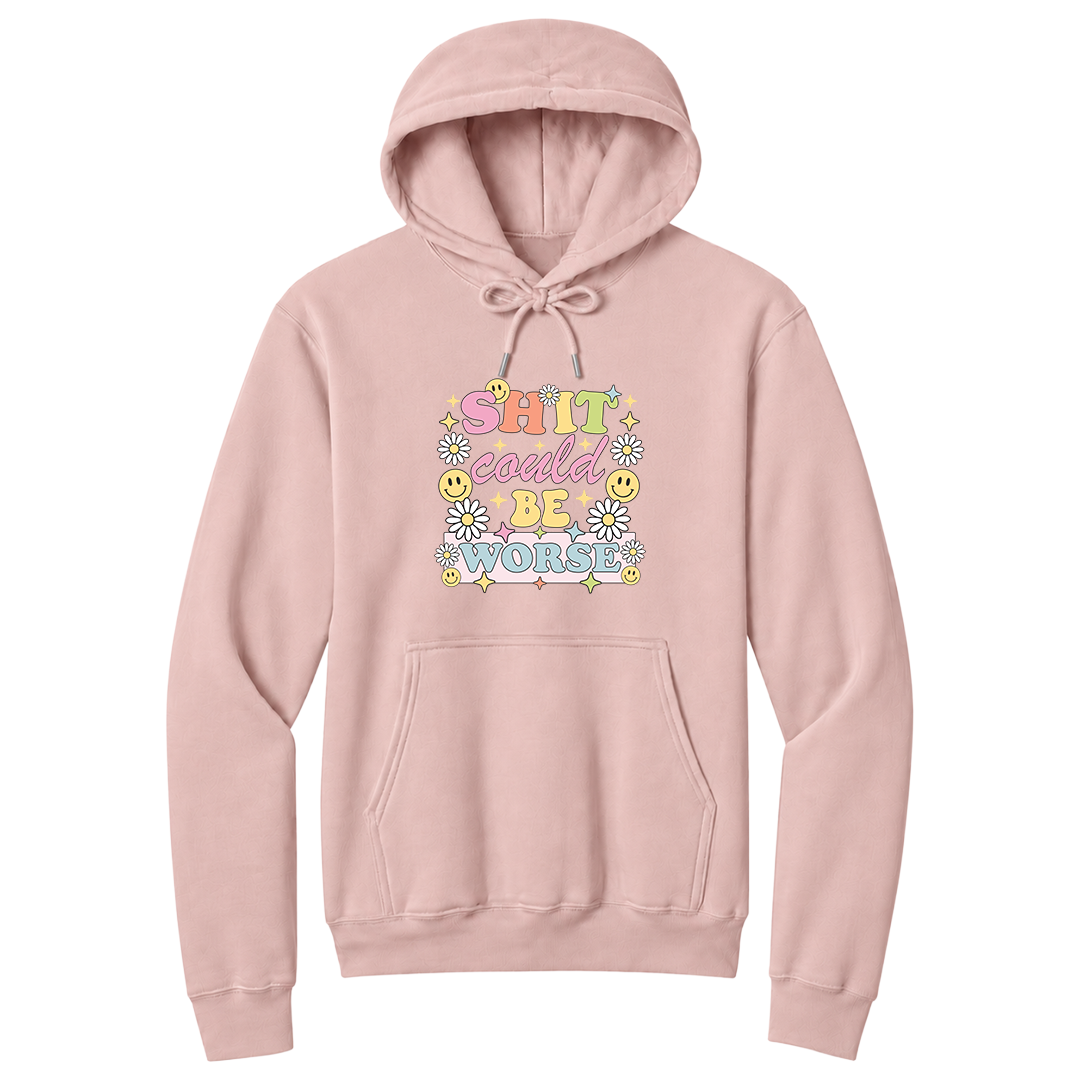 Front view of a pale blush pullover hoodie featuring a colorful retro-inspired graphic with the phrase "SHIT could BE WORSE" in playful pastel typography. The design is surrounded by white daisies, yellow smiley faces, and sparkling star accents, creating a cheerful vintage aesthetic that humorously contrasts with the sarcastic message. The drawstrings are tied into a short bow above the design for a realistic product mockup. A funny adult hoodie with bold humor, retro floral graphics, and casual style