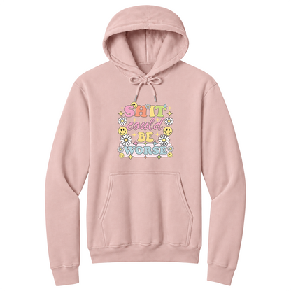 Front view of a pale blush pullover hoodie featuring a colorful retro-inspired graphic with the phrase "SHIT could BE WORSE" in playful pastel typography. The design is surrounded by white daisies, yellow smiley faces, and sparkling star accents, creating a cheerful vintage aesthetic that humorously contrasts with the sarcastic message. The drawstrings are tied into a short bow above the design for a realistic product mockup. A funny adult hoodie with bold humor, retro floral graphics, and casual style