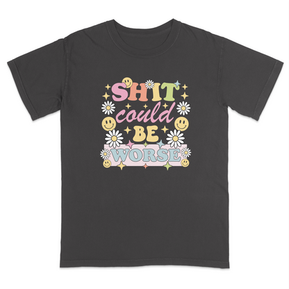 Front view of a pepper graphic t-shirt featuring a retro-inspired pastel design with the phrase "SHIT could BE WORSE" in colorful vintage lettering. The design is decorated with white daisies, yellow smiley faces, and sparkling star accents, creating a playful 70s aesthetic that contrasts with the humorous sarcastic message. A funny adult graphic tee with retro floral artwork, pastel colors, and bold statement typography for casual everyday wear.