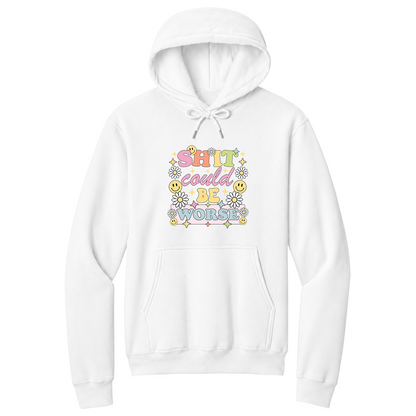 Front view of a white pullover hoodie featuring a colorful retro-inspired graphic with the phrase "SHIT could BE WORSE" in playful pastel typography. The design is surrounded by white daisies, yellow smiley faces, and sparkling star accents, creating a cheerful vintage aesthetic that humorously contrasts with the sarcastic message. The drawstrings are tied into a short bow above the design for a realistic product mockup. A funny adult hoodie with bold humor, retro floral graphics, and casual style