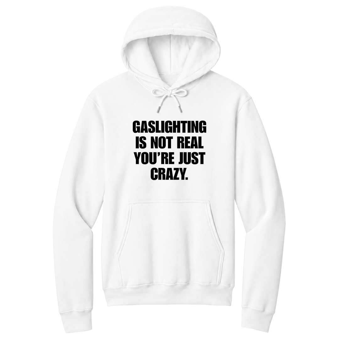 Front view of a white pullover hoodie featuring the phrase "GASLIGHTING IS NOT REAL YOU’RE JUST CRAZY." printed in bold black uppercase lettering centered on the chest. The hoodie includes a front kangaroo pocket and matching black drawstrings tied in a bow, with the minimalist black-and-white design creating a bold, sarcastic statement look.