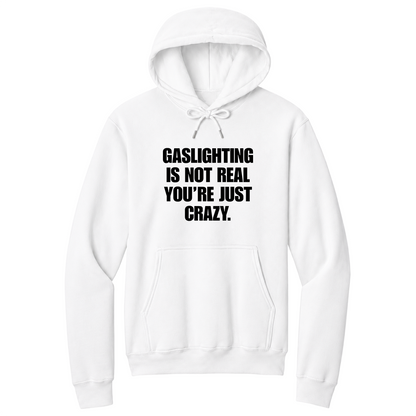 Front view of a white pullover hoodie featuring the phrase "GASLIGHTING IS NOT REAL YOU’RE JUST CRAZY." printed in bold black uppercase lettering centered on the chest. The hoodie includes a front kangaroo pocket and matching black drawstrings tied in a bow, with the minimalist black-and-white design creating a bold, sarcastic statement look.