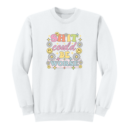 Front view of a white long sleeve graphic shirt featuring a colorful retro-inspired design with the phrase "SHIT could BE WORSE" in playful pastel typography. The graphic is surrounded by white daisies, yellow smiley faces, and sparkling star accents, creating a cheerful vintage aesthetic that humorously contrasts with the sarcastic message. A funny adult long sleeve shirt with retro floral graphics, bold humor, and casual streetwear style.