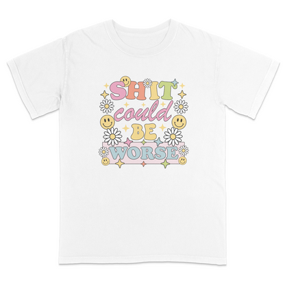 Front view of a white graphic t-shirt featuring a retro-inspired pastel design with the phrase "SHIT could BE WORSE" in colorful vintage lettering. The design is decorated with white daisies, yellow smiley faces, and sparkling star accents, creating a playful 70s aesthetic that contrasts with the humorous sarcastic message. A funny adult graphic tee with retro floral artwork, pastel colors, and bold statement typography for casual everyday wear.