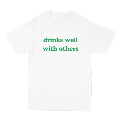 White t-shirt with green text 'drinks well with others' on a light gray background