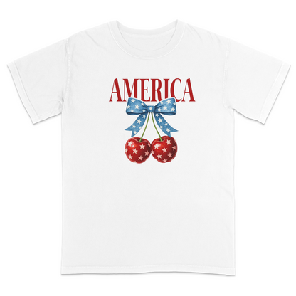 White patriotic graphic t-shirt featuring red “AMERICA” text with star-pattern cherries and a blue bow design, combining vintage Americana style with playful patriotic fashion.