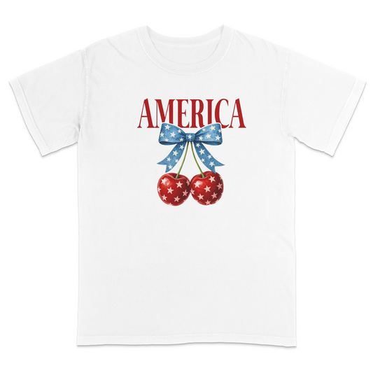 White patriotic graphic t-shirt featuring red “AMERICA” text with star-pattern cherries and a blue bow design, combining vintage Americana style with playful patriotic fashion.