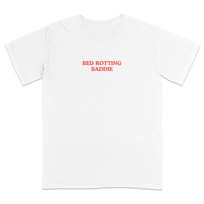 Front view of a white graphic T-shirt featuring the phrase "BED ROTTING BADDIE" in small vintage-style red lettering centered on the chest. This minimalist, trendy tee combines clean aesthetics with humorous Gen Z-inspired text for a casual everyday look.