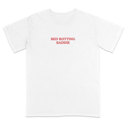 Front view of a white graphic T-shirt featuring the phrase "BED ROTTING BADDIE" in small vintage-style red lettering centered on the chest. This minimalist, trendy tee combines clean aesthetics with humorous Gen Z-inspired text for a casual everyday look.