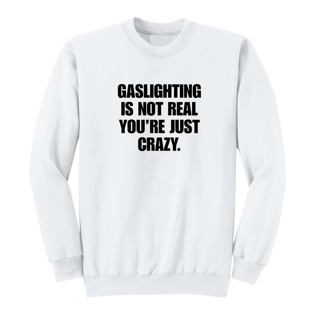 Front view of a white crewneck sweatshirt featuring the phrase "GASLIGHTING IS NOT REAL YOU’RE JUST CRAZY." printed in bold black uppercase lettering centered on the chest. The clean black-and-white design creates a minimalist statement sweatshirt with a bold, sarcastic aesthetic.