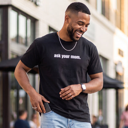 Smiling man wearing a black graphic t-shirt with white text reading “ask your mom.” paired with light-wash jeans while walking outdoors in a modern shopping district.