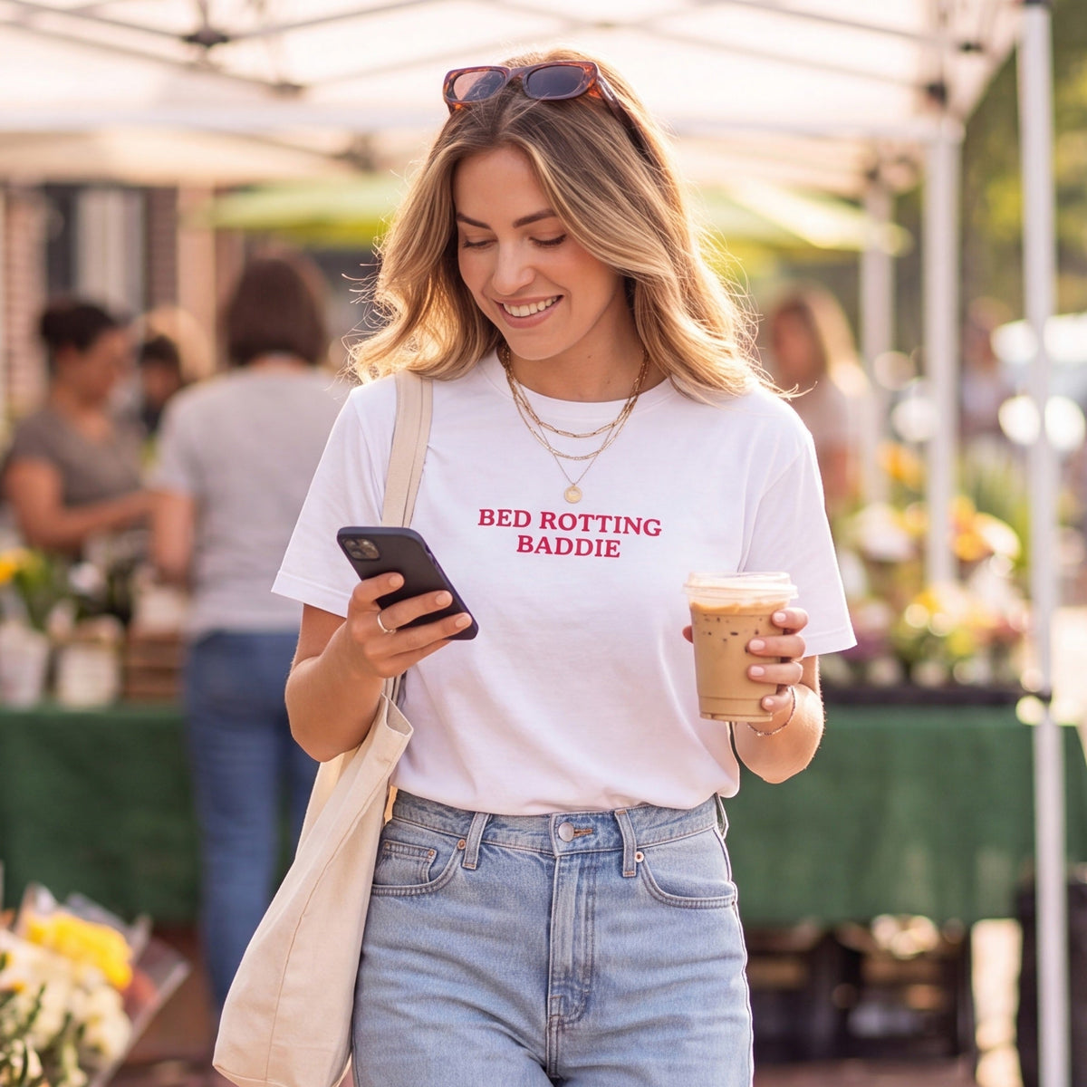 Smiling woman at an outdoor market wearing a white graphic t-shirt with red text reading “BED ROTTING BADDIE,” while looking at her phone and holding an iced coffee. She carries a canvas tote bag and wears light-wash jeans in a bright, casual lifestyle setting.