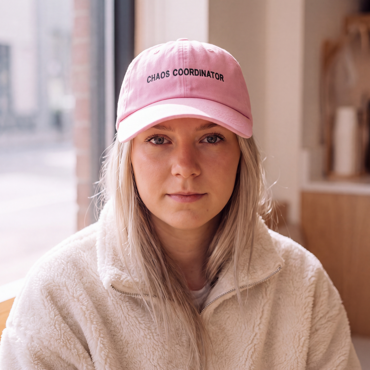 A woman wearing a light pink baseball cap with the words 'CHAOS COORDINATOR' embroidered in black on the front, paired with a cream-colored fleece jacket, photographed in a softly lit indoor setting.