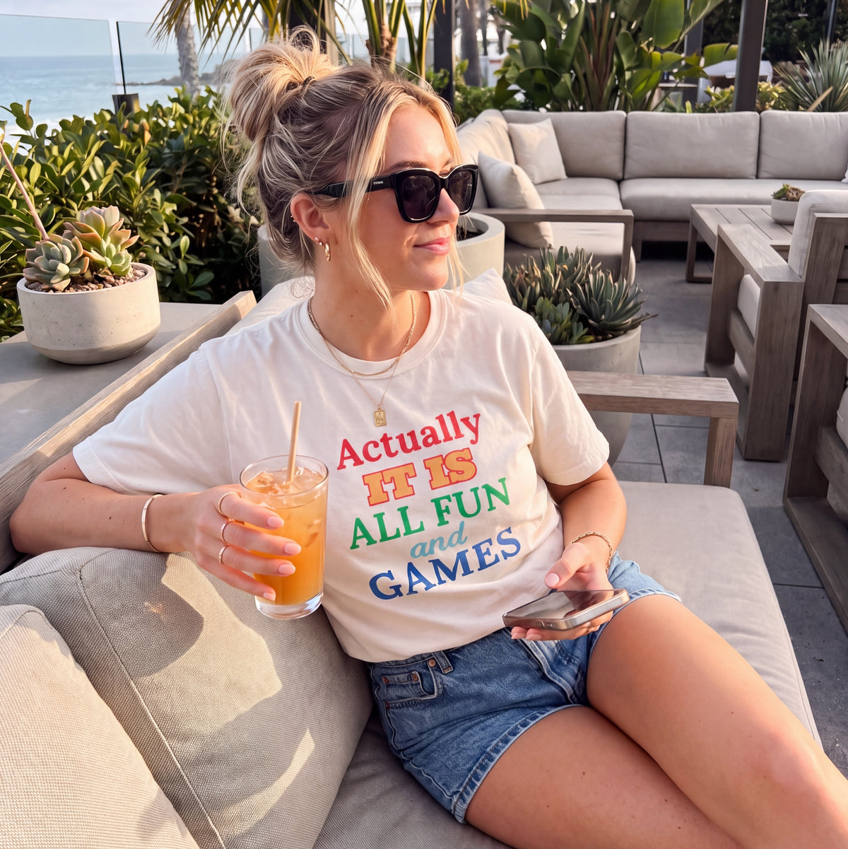 Woman in sunglasses wearing a white graphic t-shirt reading “Actually It Is All Fun and Games,” relaxing on an outdoor patio with a drink and phone.