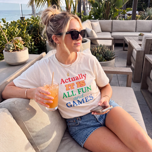 Woman in sunglasses wearing a white graphic t-shirt reading “Actually It Is All Fun and Games,” relaxing on an outdoor patio with a drink and phone.