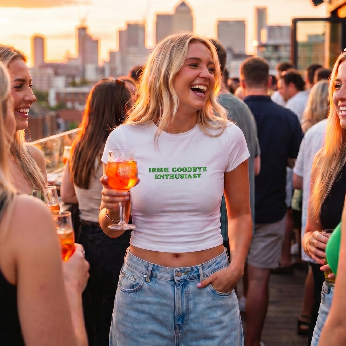 A woman laughing on a rooftop bar at sunset, wearing a white crop top with green text that reads 'IRISH GOODBYE ENTHUSIAST' and light blue jeans, holding an orange cocktail.