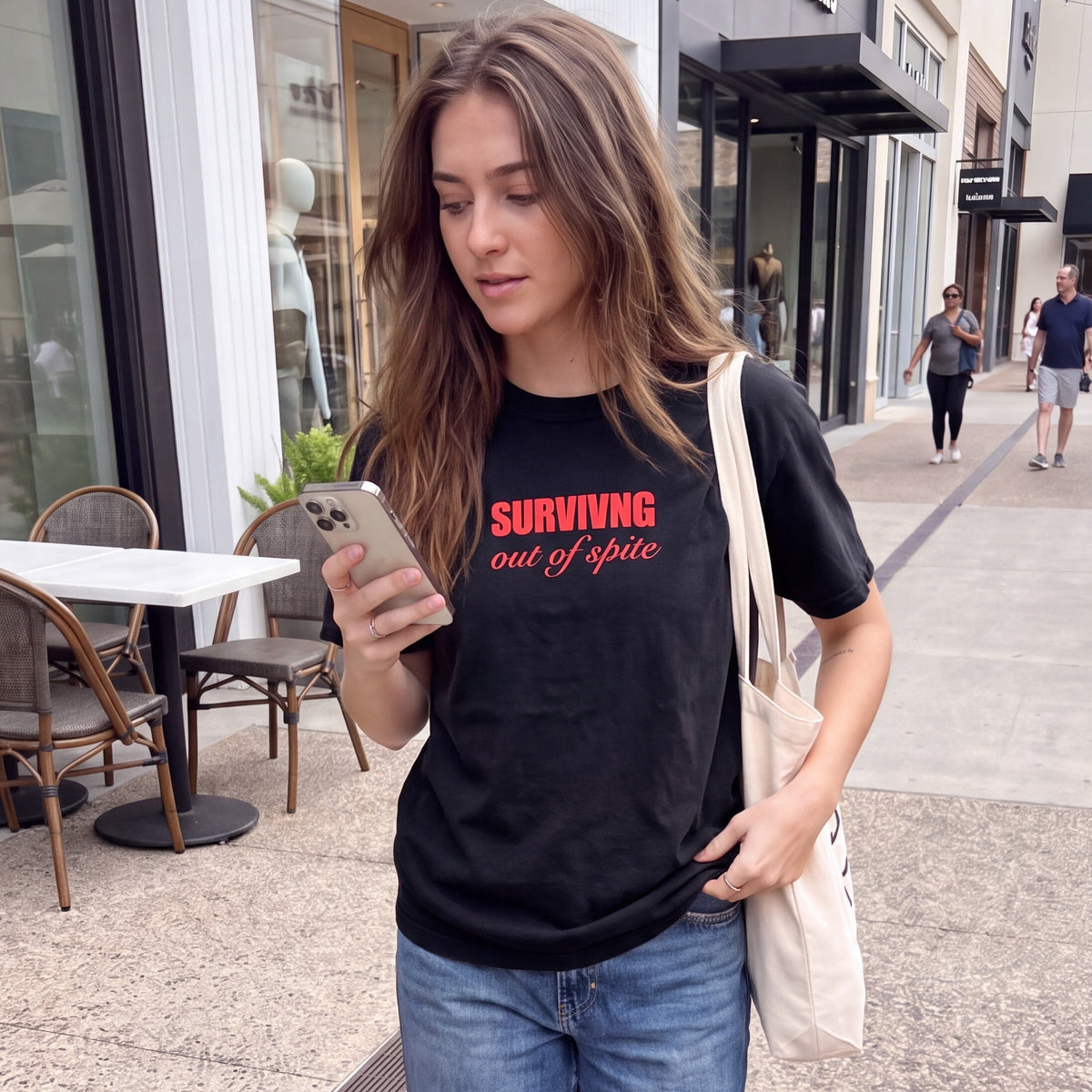 A woman walking on a city sidewalk wearing a black graphic t-shirt with red text that reads 'SURVIVING out of spite' and blue jeans.